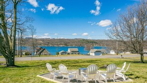 A grassy area with white Adirondack chairs and a small table, overlooking a lake with houses in the background. - Lakeview w/Pool, Hot Tub, Firepit, Gameroom, Golf! (Penn Yan)