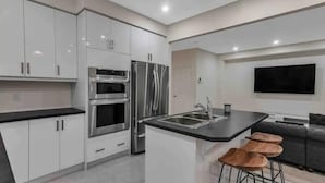A modern kitchen with stainless steel appliances, a central island with stools, and a grey sofa in the adjacent living area.
