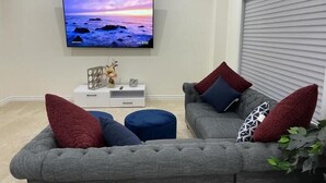 A living room with a grey sofa, blue ottoman, and a flat-screen TV displaying a sunset over the ocean.