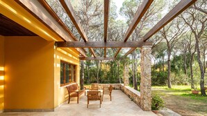 An outdoor patio with wooden furniture, a stone pillar, and a view of a forested area.