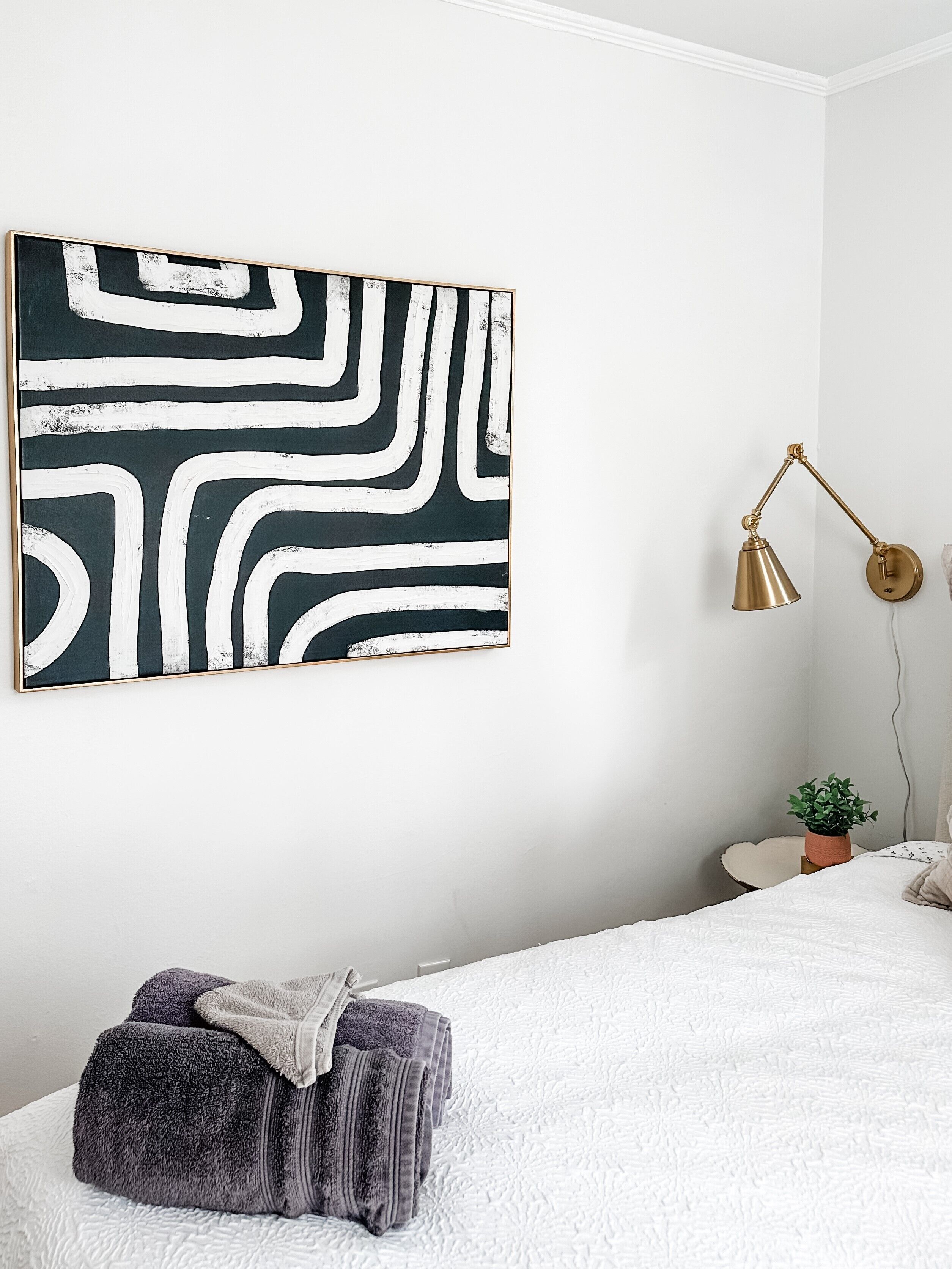 Une chambre à coucher avec un lit, une lampe fixée au mur, une plante en pot et un grand tableau abstrait.