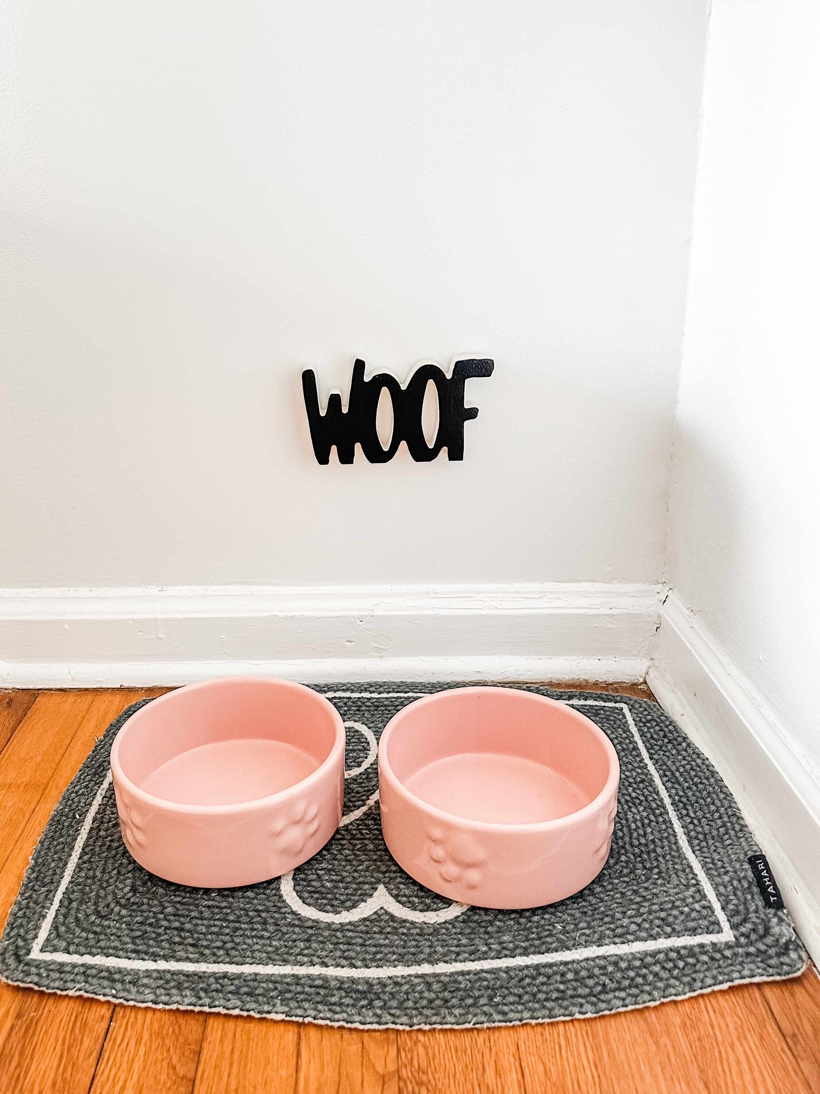 A corner of a room with a white wall, a black 'WOOF' sign, and two pink pet bowls on a grey mat.