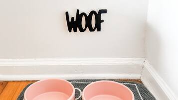 A corner of a room with a white wall, a black 'WOOF' sign, and two pink pet bowls on a grey mat.