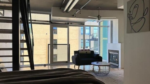 A modern hotel room with a large bed, a small seating area, a fireplace, and a view of the city through large windows.