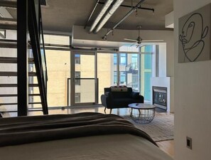 Una habitación de hotel moderna con una cama grande, una pequeña zona de estar, una chimenea y vistas a la ciudad a través de amplios ventanales.