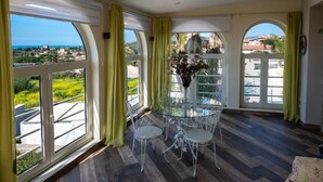 A room with a glass table and chairs, large windows with curtains, and a view of greenery and buildings.