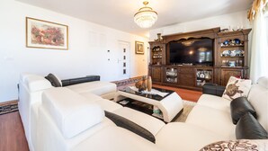 Ein Wohnzimmer mit einem weißen Sofagarnitur, einem Couchtisch mit Dekoration, einem Fernseher auf einer Holzkommode und einer Kristallleuchter.
