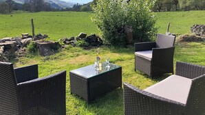 An outdoor seating area with wicker furniture and a table, set against a backdrop of a green field and distant mountains.