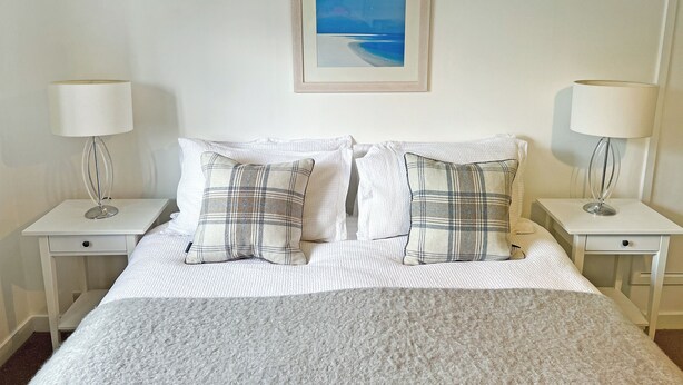 A neatly made bed with white bedding and two plaid pillows, flanked by two bedside tables with lamps and a framed picture above the bed.