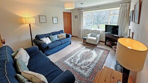 A living room with a blue sofa, a small armchair, a TV stand, and a large window with curtains.