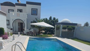A swimming pool with a tiled deck, a covered seating area, and a building with a dome and arches in the background.