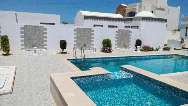A swimming pool area with a clear blue pool, surrounded by white walls and a few potted plants.