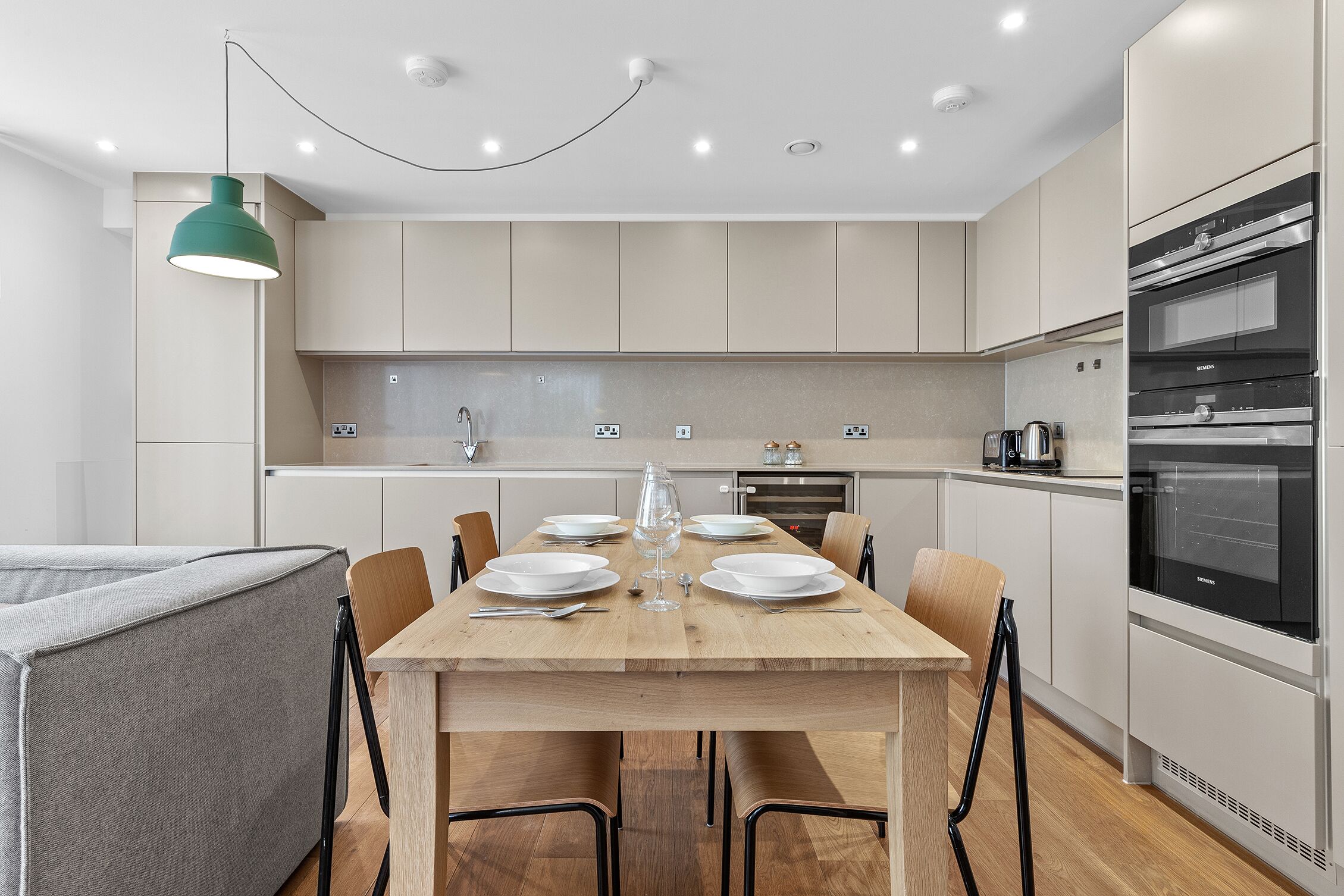 A modern kitchen with a wooden dining table set for four, a built-in oven, and a pendant light.