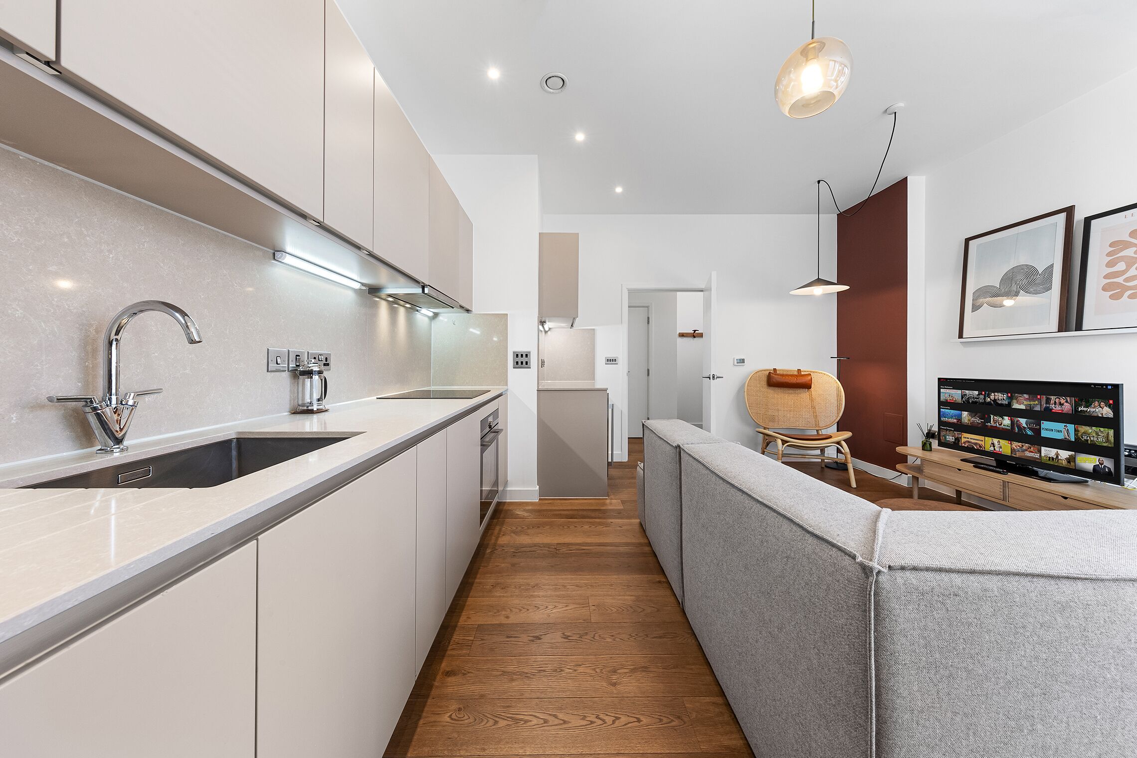 A modern kitchen with a built-in sink, a wooden floor, and a grey sofa in the living area.