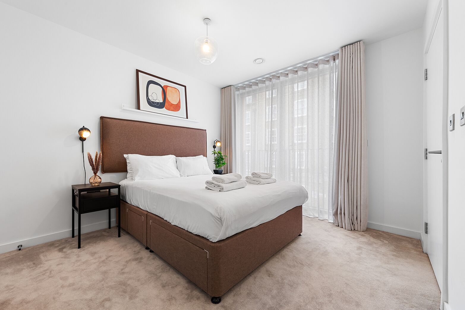 A modern bedroom with a large bed, a bedside table with a lamp, a framed picture, and sheer curtains.
