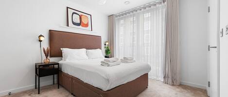 A modern bedroom with a large bed, a bedside table with a lamp, a framed picture, and sheer curtains.