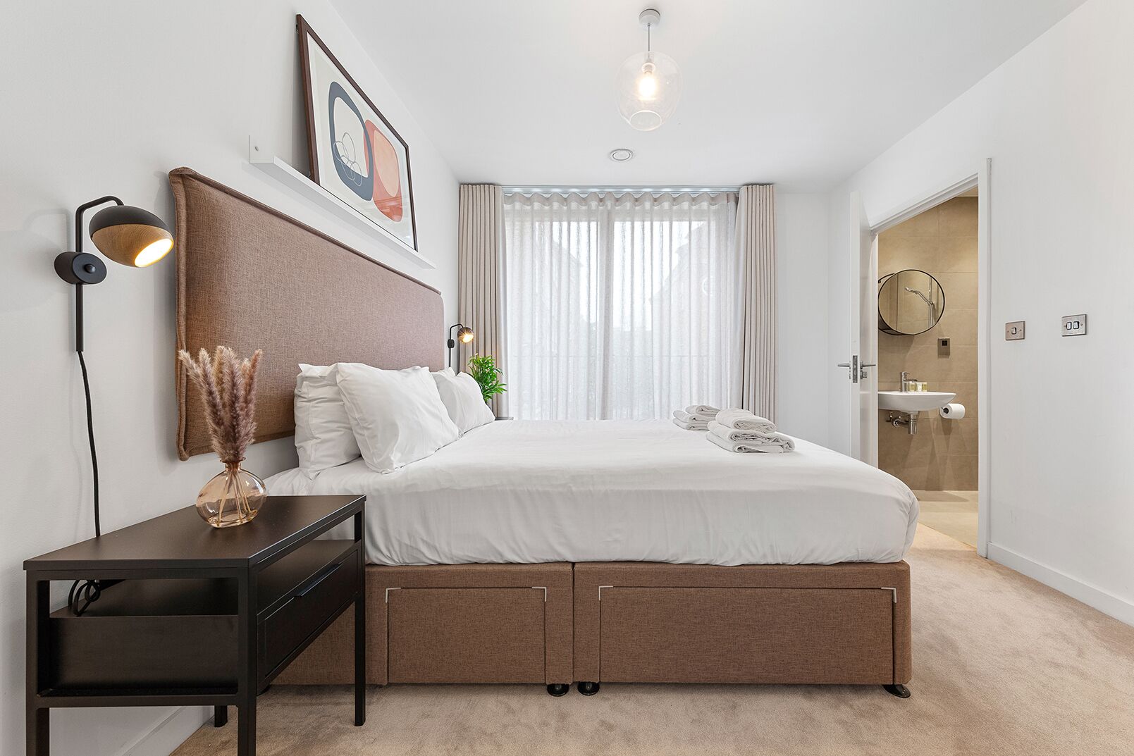 Un dormitorio moderno con cama, mesita de noche, lámpara y vistas al baño.