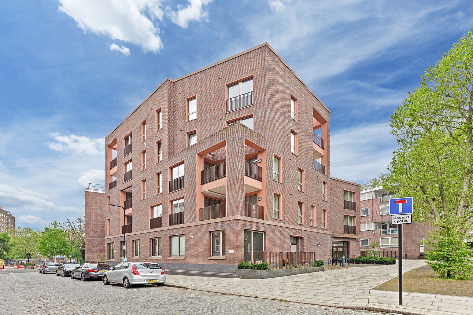 A multi-story brick building with balconies, a parking area with cars, and a clear blue sky.