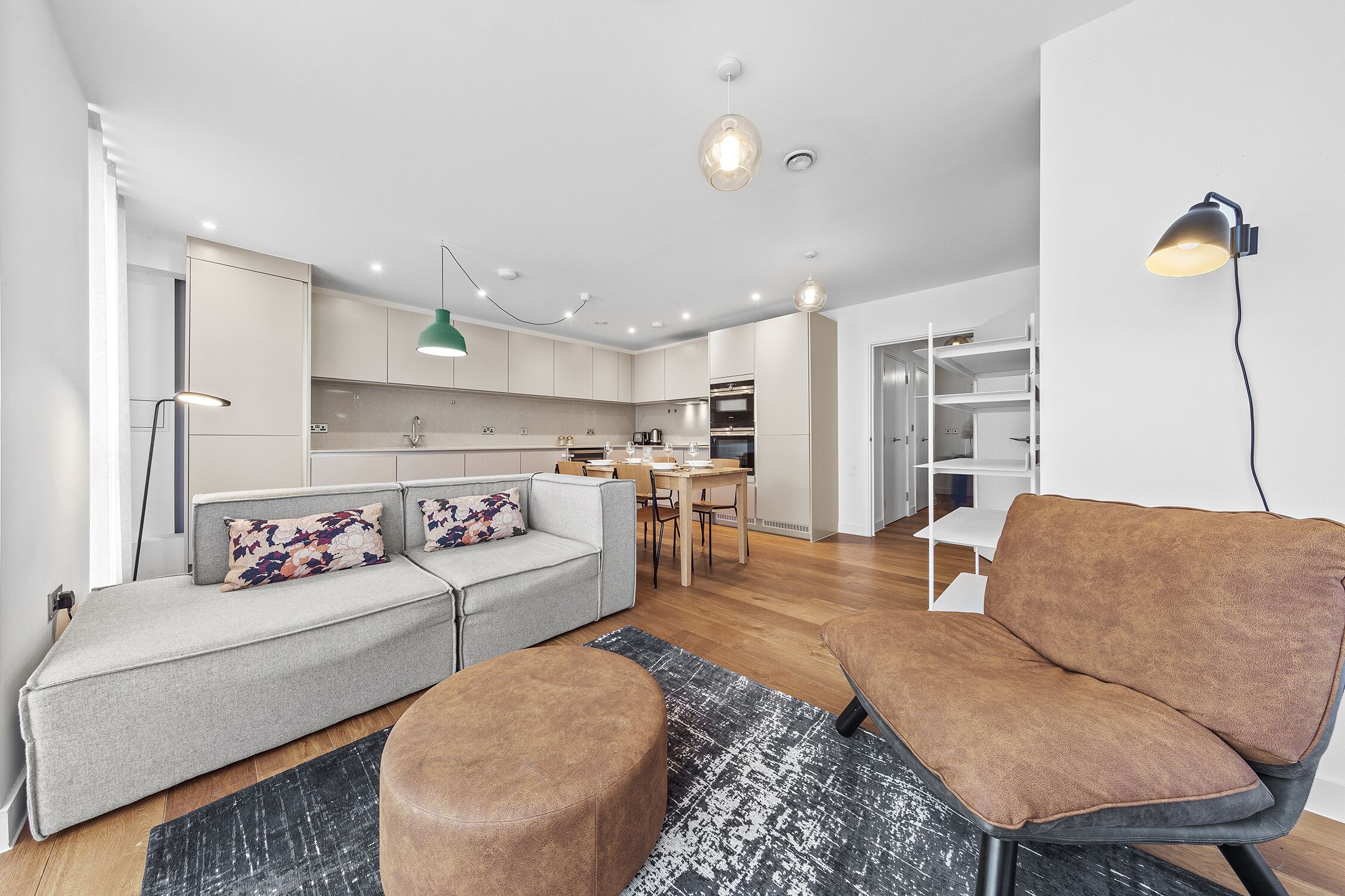 A modern living room with a grey sofa, brown armchair, and ottoman, a dining area with a table and chairs, and a kitchen with white cabinets and a hanging light.