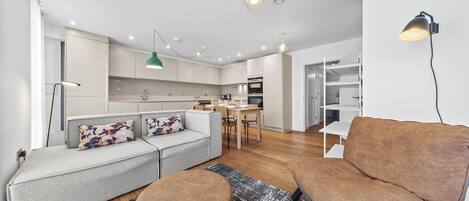 A modern living room with a grey sofa, brown armchair, and ottoman, a dining area with a table and chairs, and a kitchen with white cabinets and a hanging light.