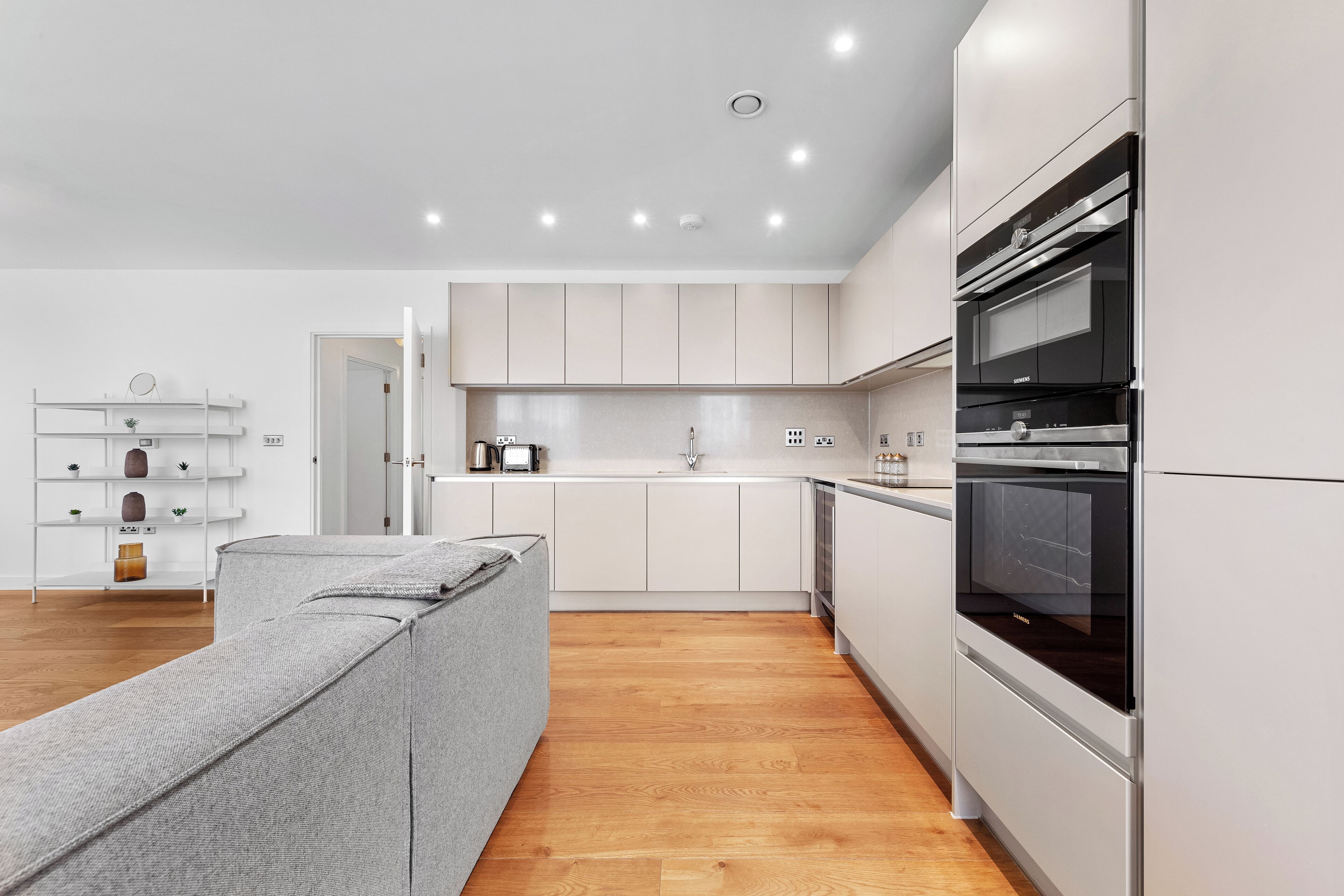 Una cocina moderna con armarios blancos, horno empotrado y un sofá gris en la sala.