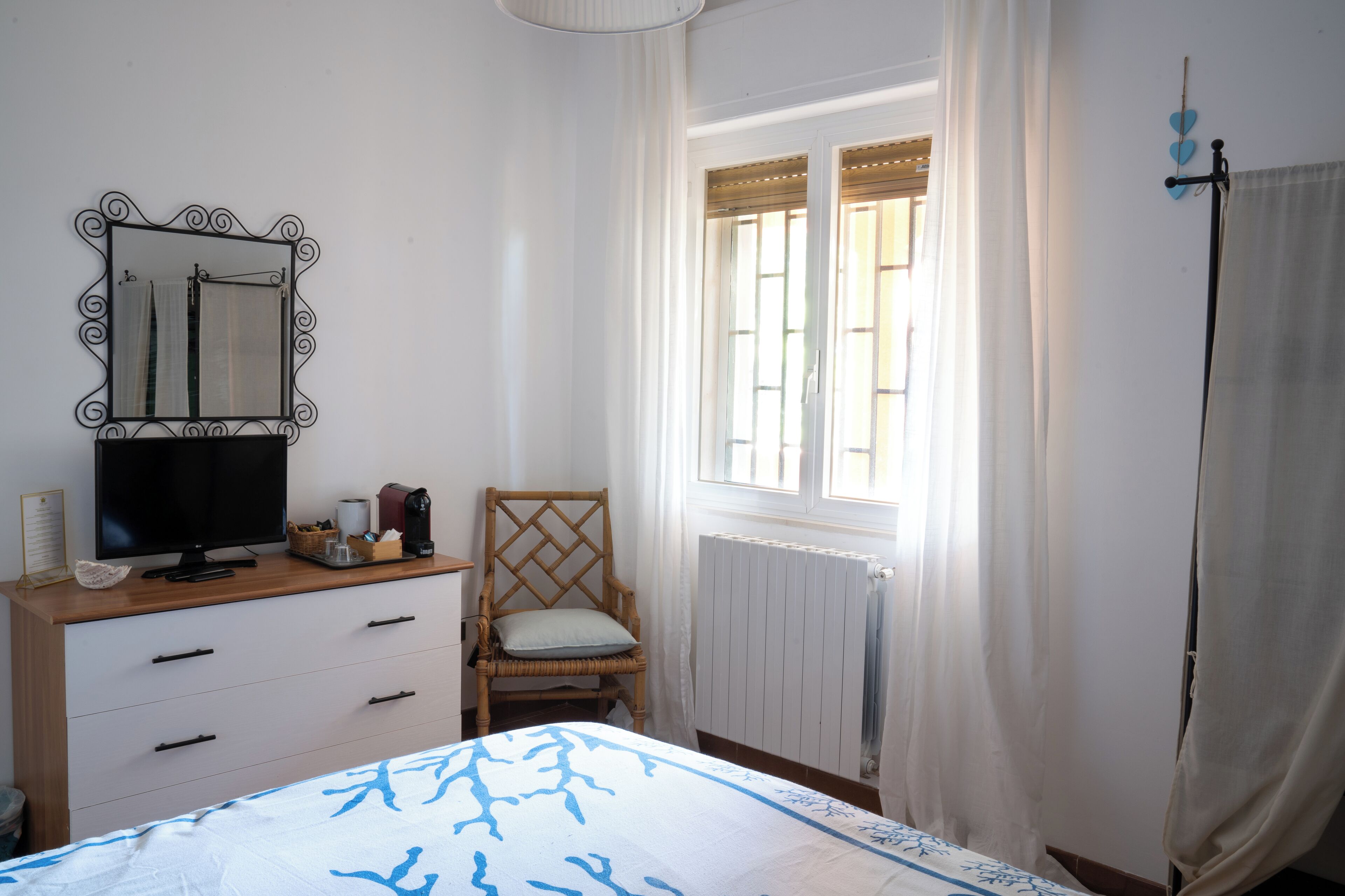 Une chambre à coucher comprenant un lit, une commode, une chaise, un téléviseur, une fenêtre avec des rideaux et un radiateur.