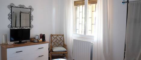 Une chambre à coucher comprenant un lit, une commode, une chaise, un téléviseur, une fenêtre avec des rideaux et un radiateur.