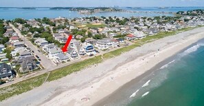 An aerial view of a coastal residential area with houses along the beach, a clear view of the ocean, and a red arrow pointing to a specific location.