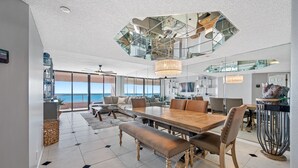 Un vaste espace dédié à la salle à manger, avec une grande table en bois et des chaises en cuir, un plafond haut orné d’un miroir aux motifs géométriques, et une vue sur l’océan grâce à de grandes fenêtres.