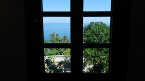 Uma janela com quatro painéis, cada um com uma vista diferente: céu azul límpido, mar distante, árvore verde densa e um jardim com plantas e um muro de pedra.