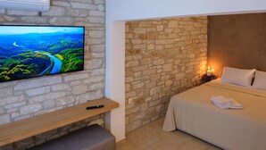 Quarto com parede de pedra, uma cama, mesa de cabeceira com um controle remoto e uma TV na parede exibindo uma paisagem cênica.