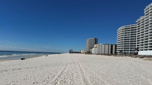 Une vaste plage de sable fin parsemée d’empreintes, un ciel bleu azur et des bâtiments qui se dessinent à l’horizon.