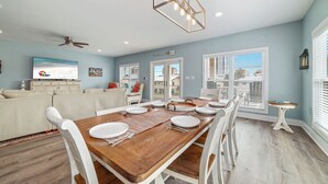 Un coin repas avec une table en bois dressée pour quatre personnes, des chaises blanches et un ventilateur de plafond.