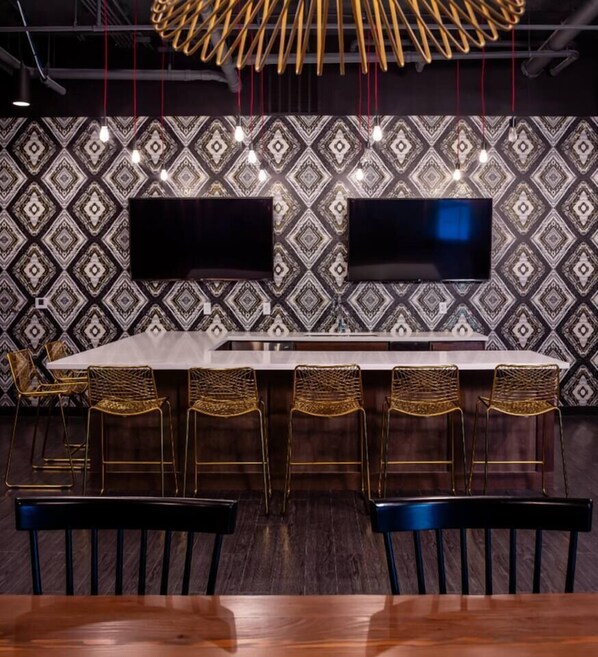 Zona de bar con una mesa larga, cuatro taburetes y dos televisores de pantalla plana montados en una pared decorada.
