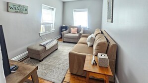 A living room with a grey sofa, a grey armchair, a small ottoman, a wooden coffee table, and a side table with a tissue box. There is a wall-mounted shelf with books and a decorative sign. - Alice Pet-Friendly Downtown Newport Rental with Parking  (Newport)