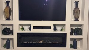 Un salon doté d’un grand téléviseur à écran plat fixé à un mur blanc, de part et d’autre duquel se trouvent des étagères ornées d’objets décoratifs, et une cheminée en dessous.