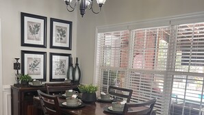Un coin repas avec une table en bois dressée pour quatre personnes, entourée de chaises. Les murs sont ornés d’illustrations botaniques encadrées, un lustre est suspendu au plafond et une fenêtre est dotée de stores.