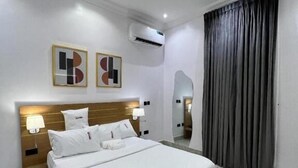 Una habitación de hotel moderna con una cama grande, cabecera con iluminación integrada, mesita de noche con lámpara, un espejo amplio y cortina gris.