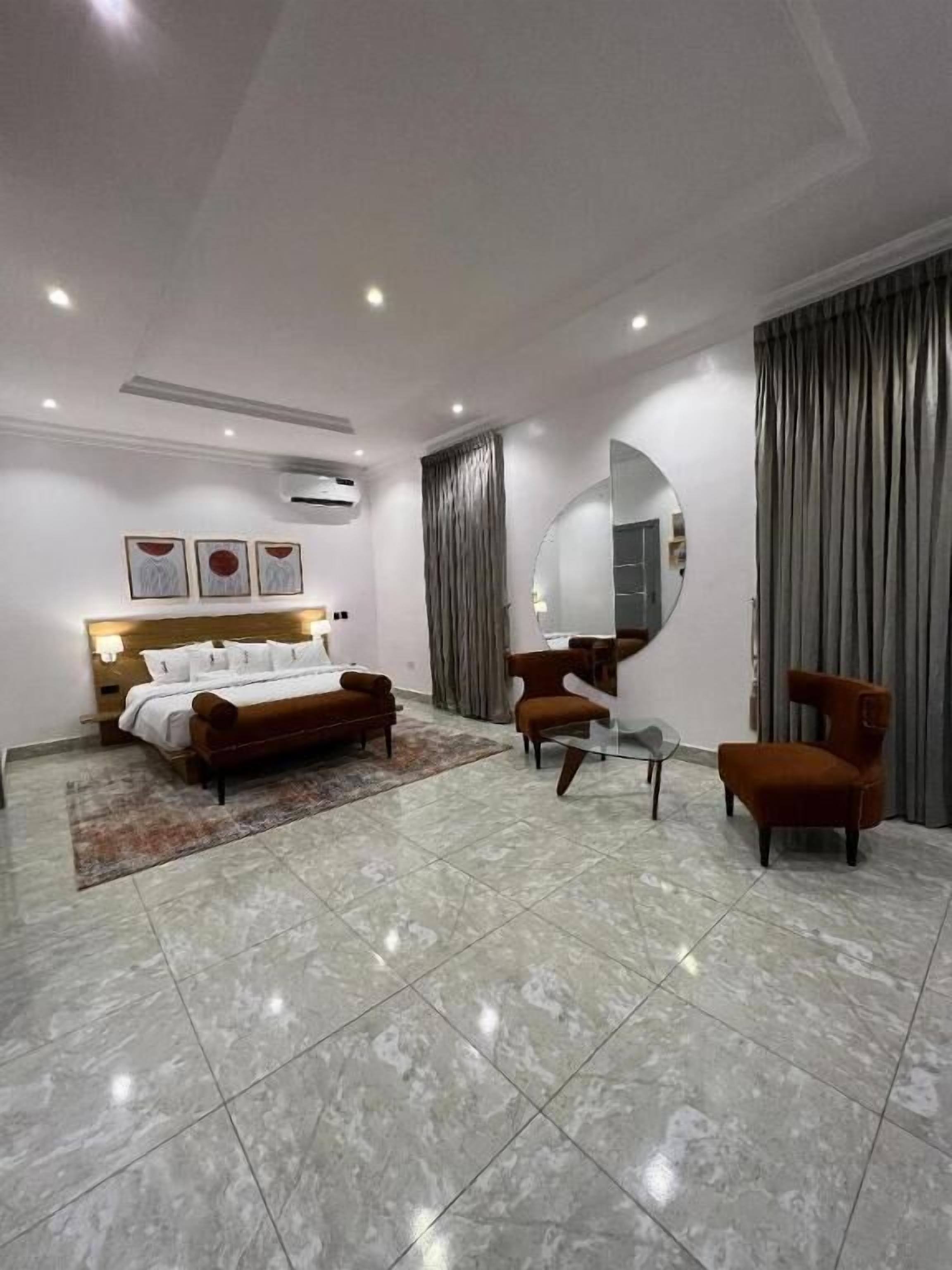 A modern hotel room with a bed, two armchairs, a round mirror, and a large window with curtains.