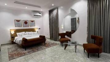 A modern hotel room with a bed, two armchairs, a round mirror, and a large window with curtains.
