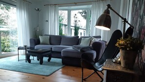 Een woonkamer met een grijze bank, een zwarte salontafel en een zwarte fauteuil. Er staat een lamp op een bijzettafeltje en een raam met gordijnen.