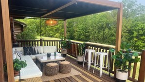 A covered deck with a sofa, table, and chairs, surrounded by potted plants and a wooden railing. - Private Cabin Resort l Pool • Hot Tub • Sauna Wanderlust Cabin (Lodi)