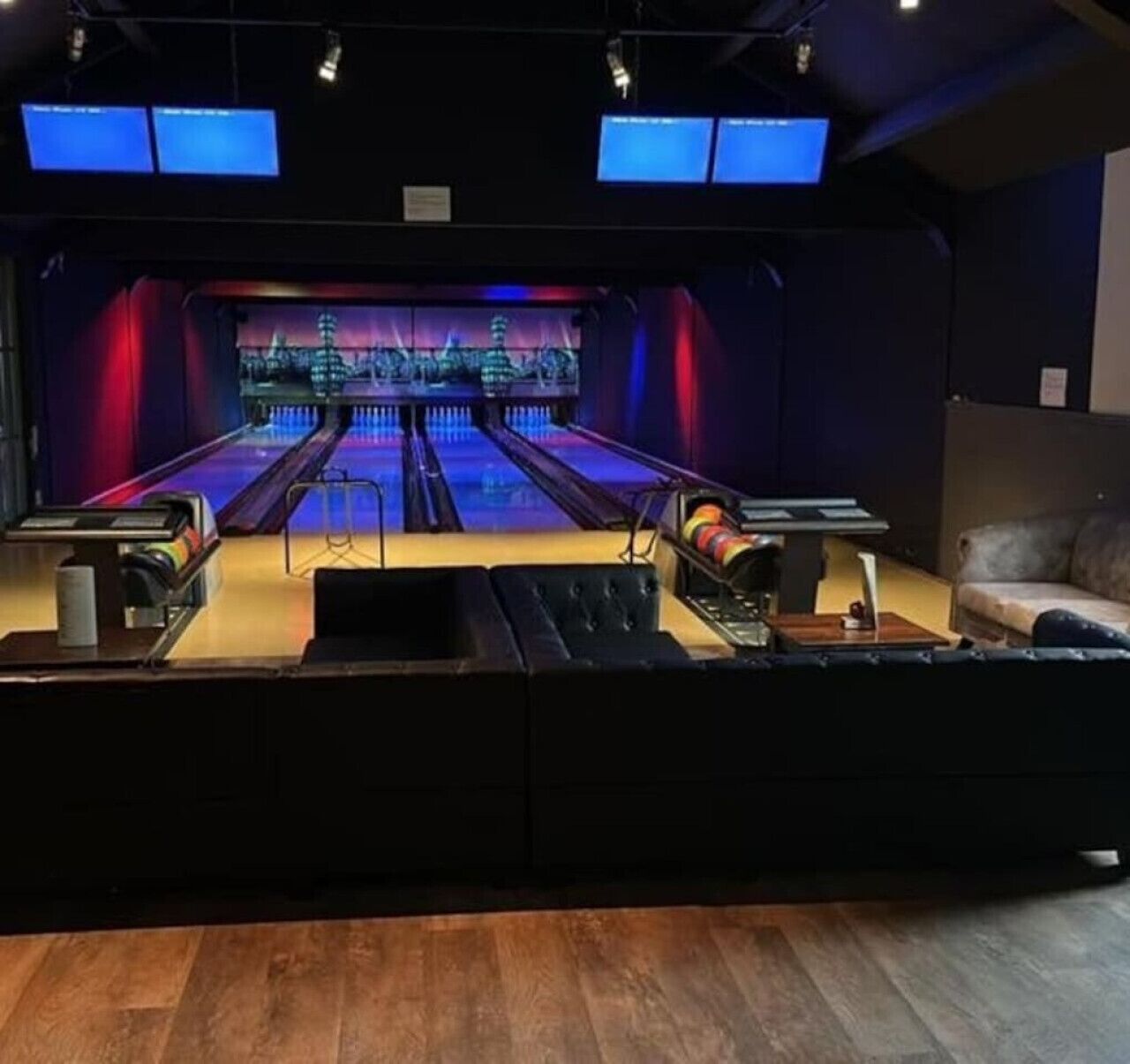 A bowling alley with a wooden floor, two bowling lanes, and a couch in the foreground.