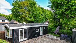 Un piccolo chalet in legno nero con porta e finestre in vetro, circondato da un patio in ghiaia e rigogliosa vegetazione.