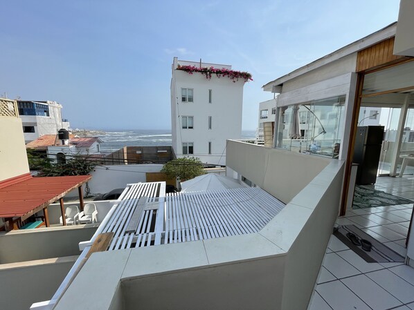 A rooftop terrace with a view of the sea, a modern building with a balcony, and a glass door leading to an indoor area.