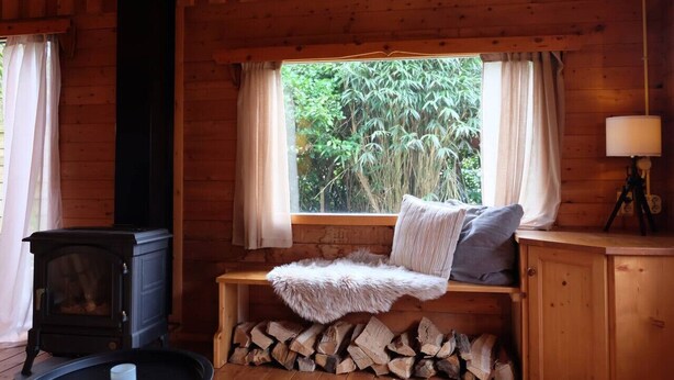 A cozy cabin interior with a wood-burning stove, a bench with a fur throw, a window with curtains, and a tripod lamp.