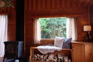 A cozy cabin interior with a wood-burning stove, a bench with a fur throw, a window with curtains, and a tripod lamp.