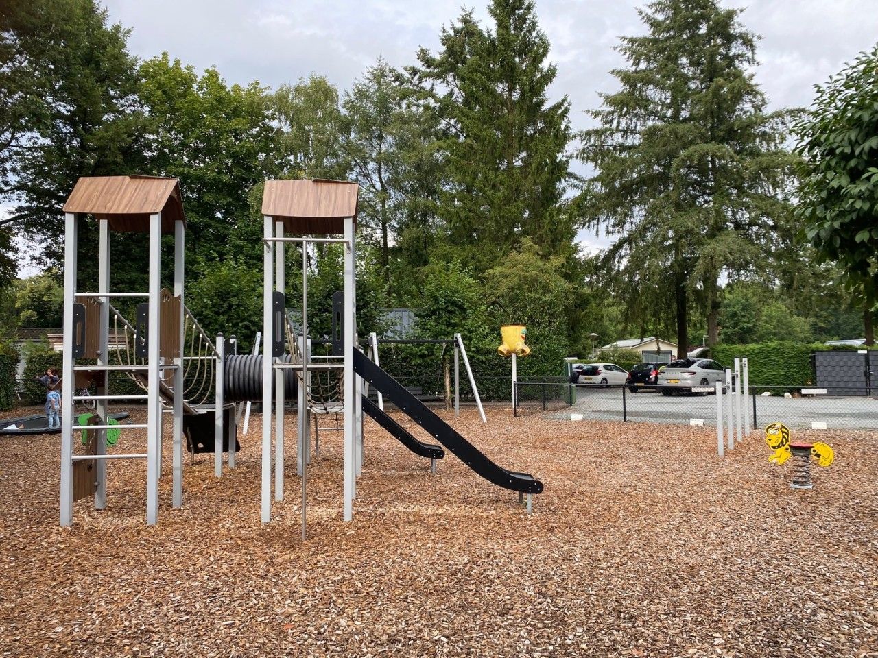 A playground with a slide and climbing structures, surrounded by trees and a parking area.