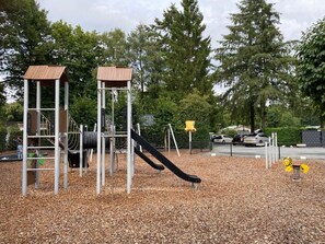A playground with a slide and climbing structures, surrounded by trees and a parking area.