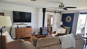 A living room with a large flat-screen TV, a ceiling fan, a fireplace, and a view of the outdoors. - Year Around View Lake Tenkiller (Vian)